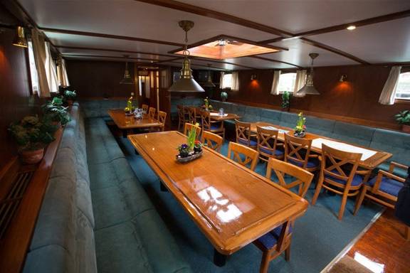 Salon met houten eettafels en groene zitbanken aan boord van een zeilschip